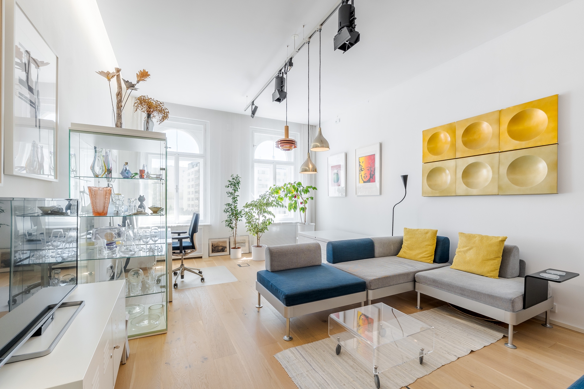 Bright living room with a modular gray and blue sofa, yellow cushions, glass coffee table, and gold wall art.