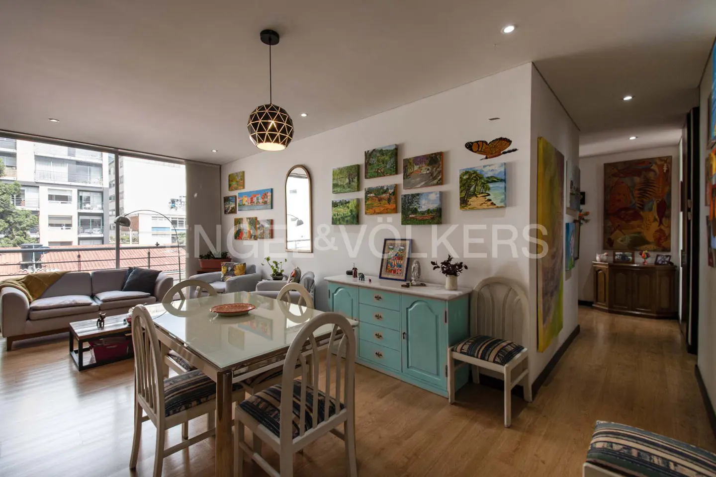 Bright living and dining area with a white table, chairs, and a blue sideboard. Art adorns the walls, and a large window offers a city view.