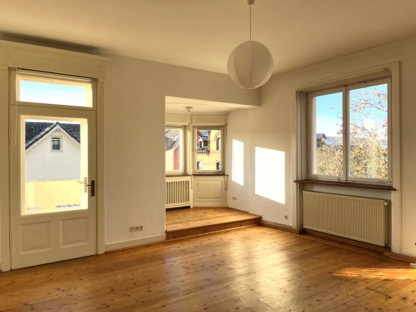 Bright, empty room with wood floors, white walls, and a paper lantern. A doorway and window offer views of houses and trees.
