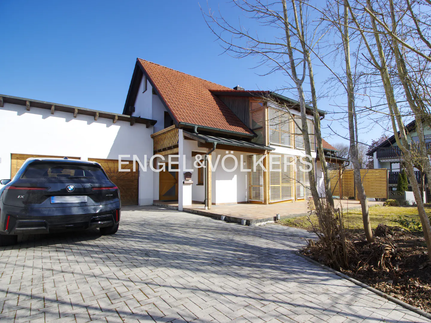 Exterior view of a two-story white house with a red tile roof and a black BMW parked in the driveway. The Engel & Völkers logo is visible.