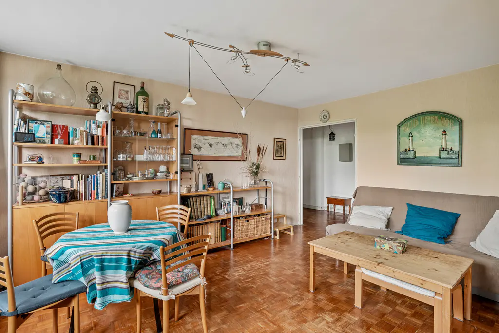 Cozy living room with parquet floors, bookshelves, and a dining table with blue striped tablecloth. A tan sofa with blue pillows sits near a lighthouse painting.