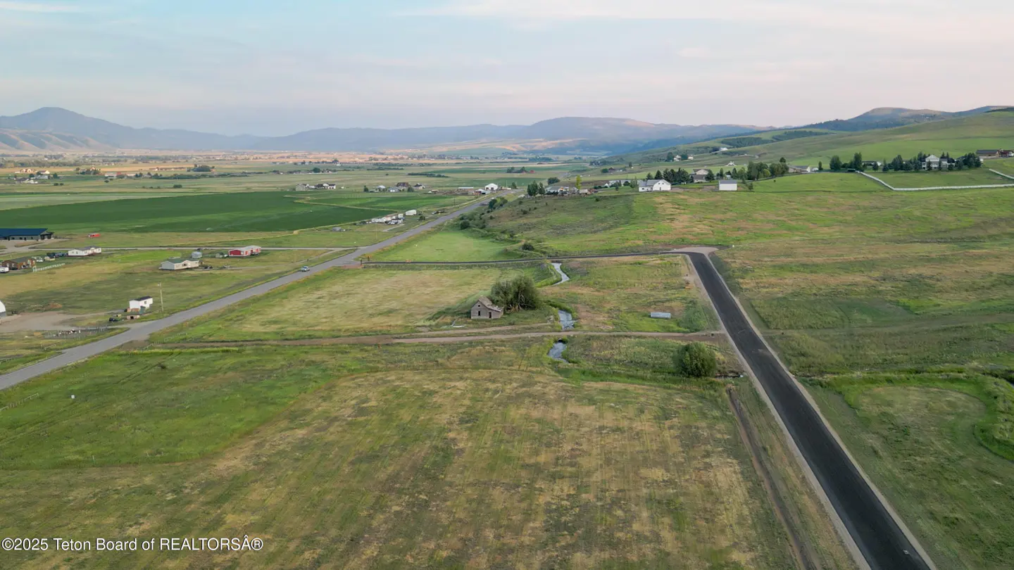 Aerial view of a green, grassy field with roads, a stream, and a small, weathered building. Mountains are visible in the background.