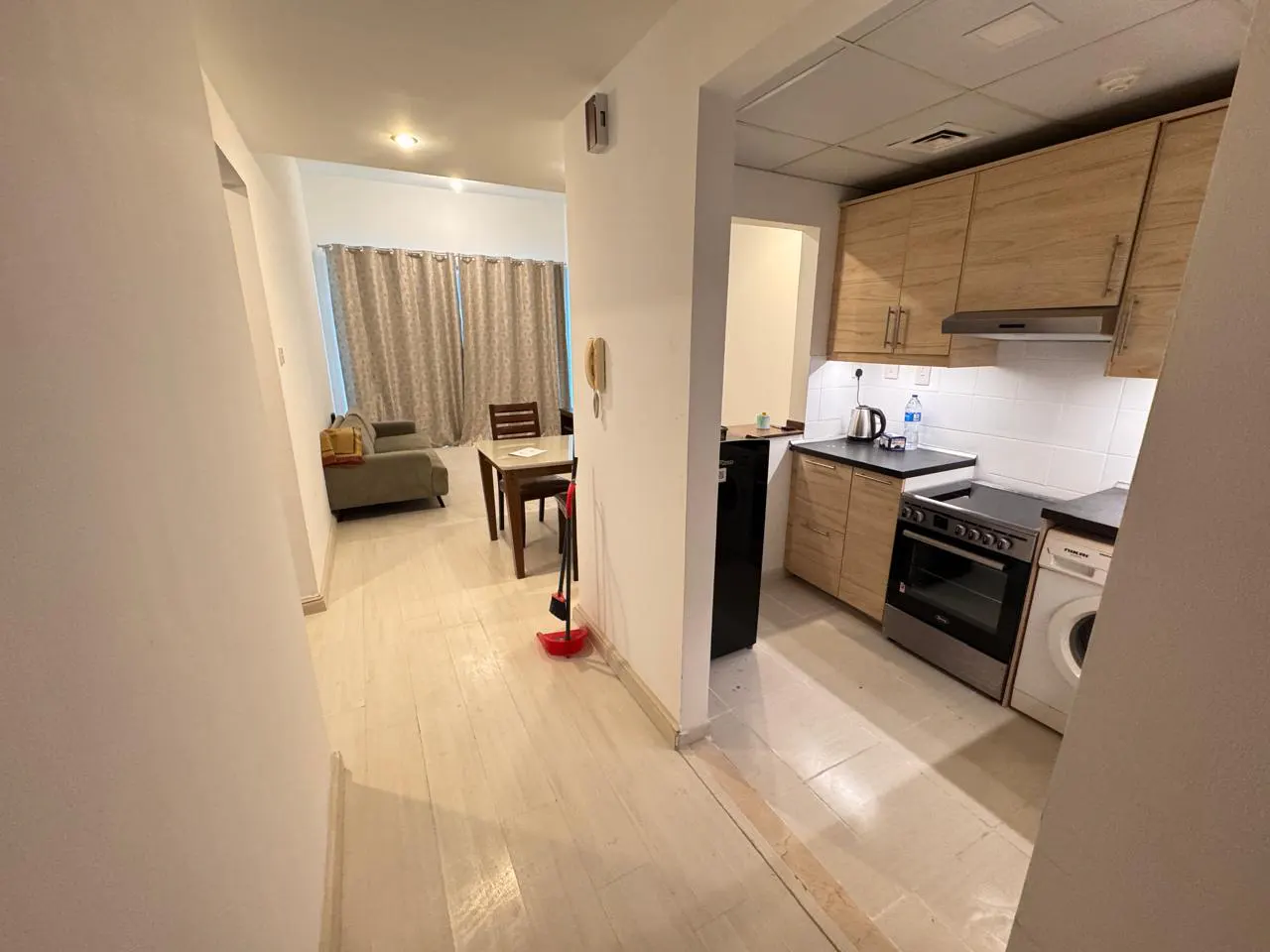 A view of a modern apartment with a kitchen, dining area, and living room. The kitchen has wooden cabinets and appliances. The living room has a green sofa and curtains.