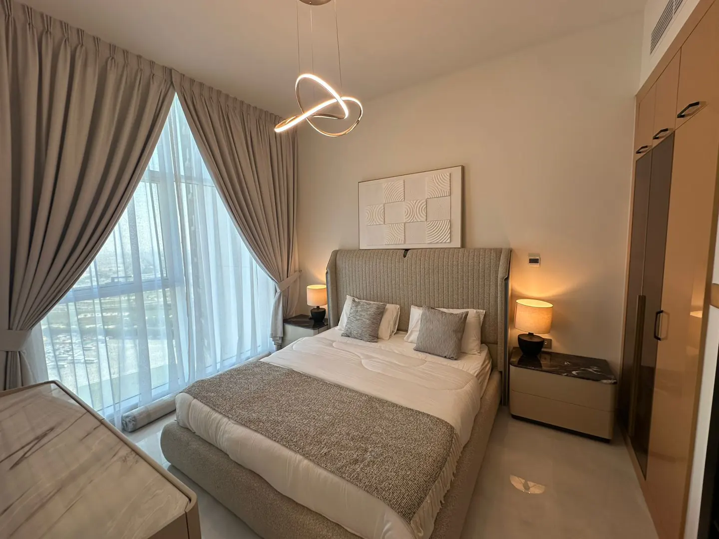 A modern bedroom with a large bed, beige headboard, and neutral-toned bedding. A window with sheer curtains provides natural light. A unique light fixture hangs above.
