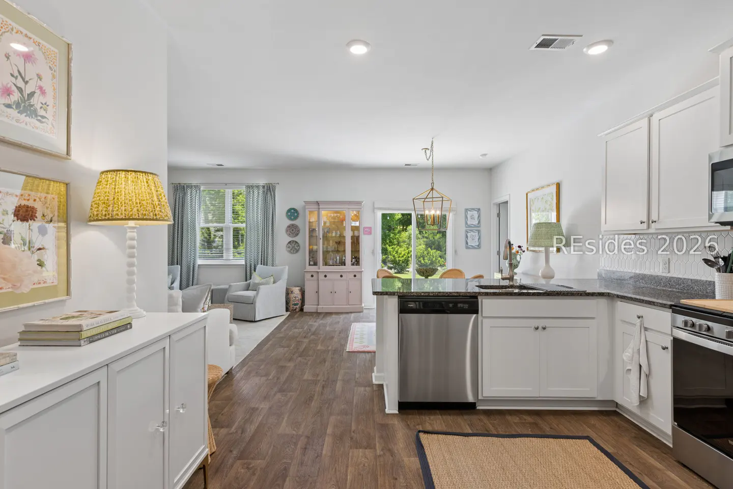 Open-concept living space with white cabinets, stainless steel appliances, and wood floors. A living room is visible in the background.