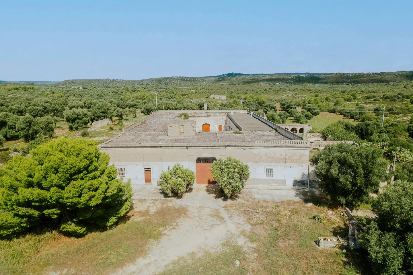 Aerial view of a large, one-story, white farmhouse with a flat roof and orange doors, surrounded by trees and fields.