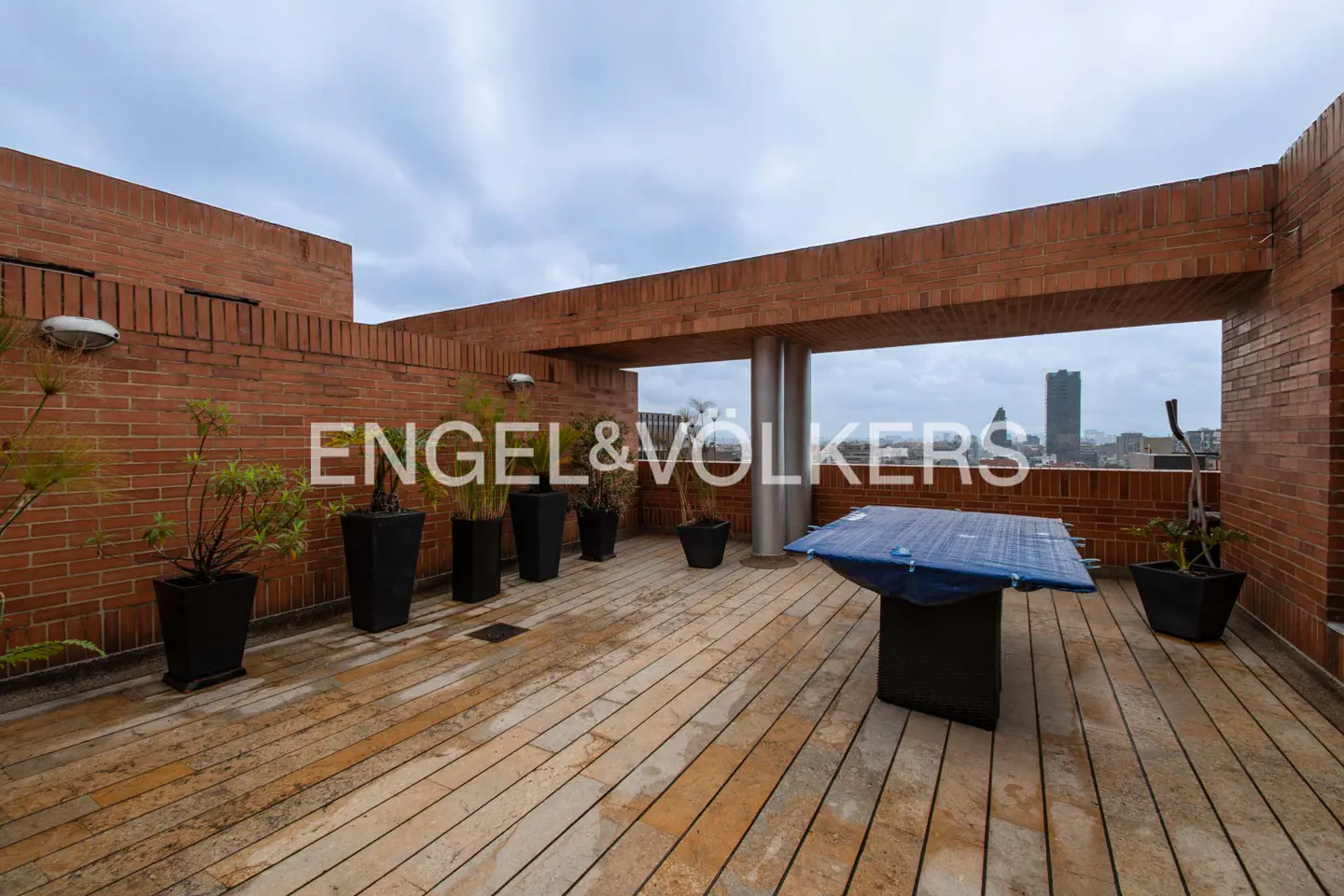 Rooftop patio with brick walls, potted plants, and a table covered with a blue tarp. City skyline visible in the background.