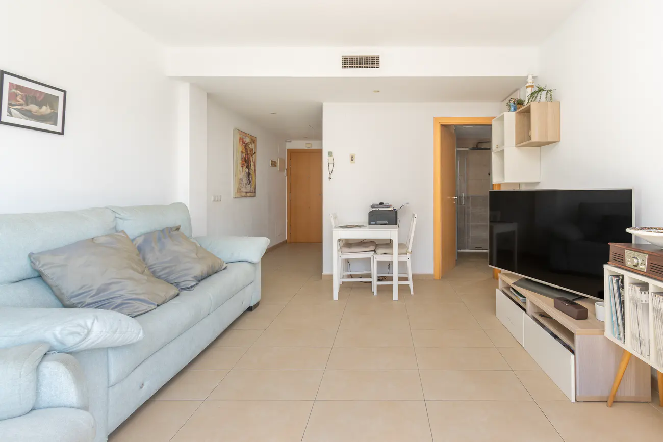 Bright living room with a light blue sofa, white walls, and beige tile flooring. A small white table and chairs sit near a doorway. A TV and shelving unit are on the right.