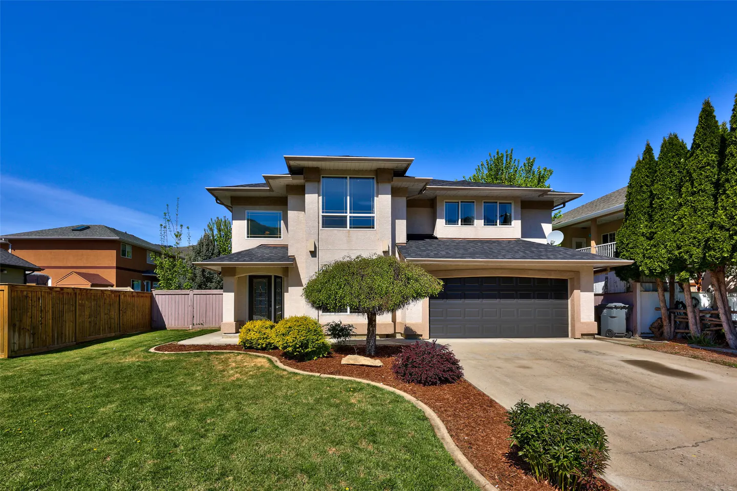 Two-story beige house with a dark gray garage door, green lawn, and blue sky.