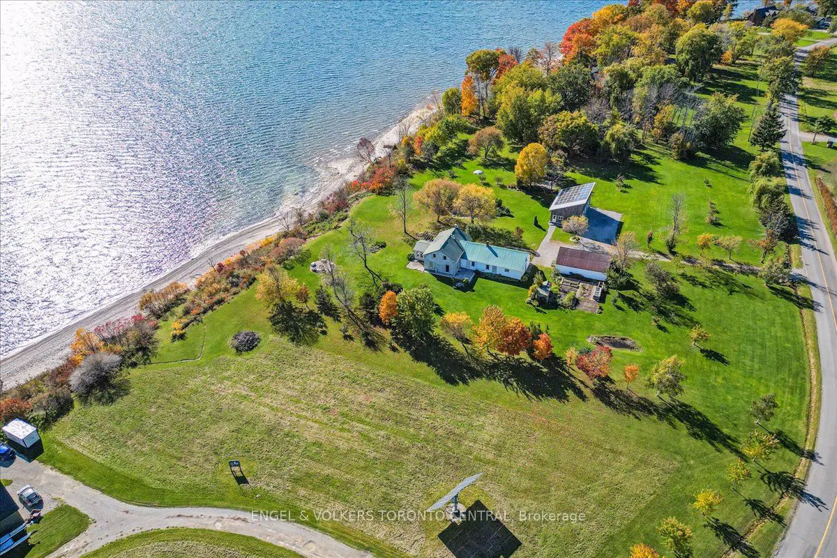 Aerial view of a waterfront property with a house, barn, and green lawn. Trees in autumn colors line the shore and a road borders the property.