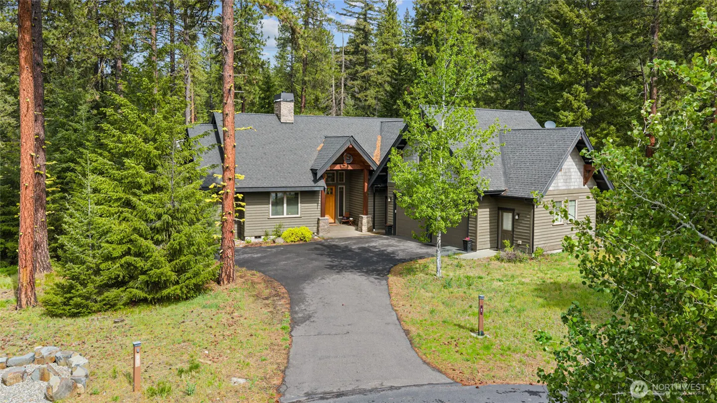 Exterior view of a gray house with a dark roof, surrounded by green trees and a paved driveway.