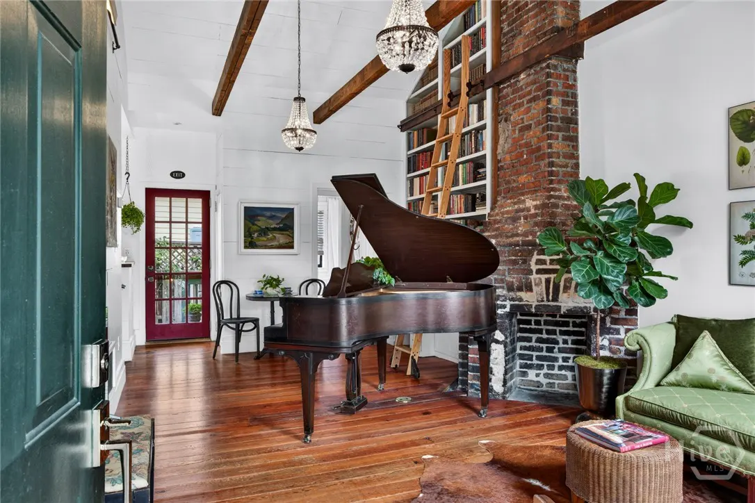 Interior view of a living room with a grand piano, brick fireplace, and wooden floors. Chandeliers hang from the ceiling.