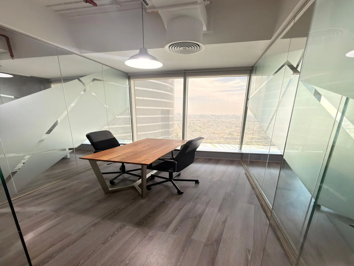 Modern office meeting room with a wooden table, two black chairs, glass walls, and a high-rise city view through windows.
