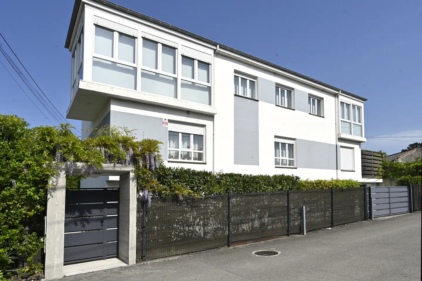 Exterior view of a modern two-story house with white and gray facade, surrounded by a fence and greenery.