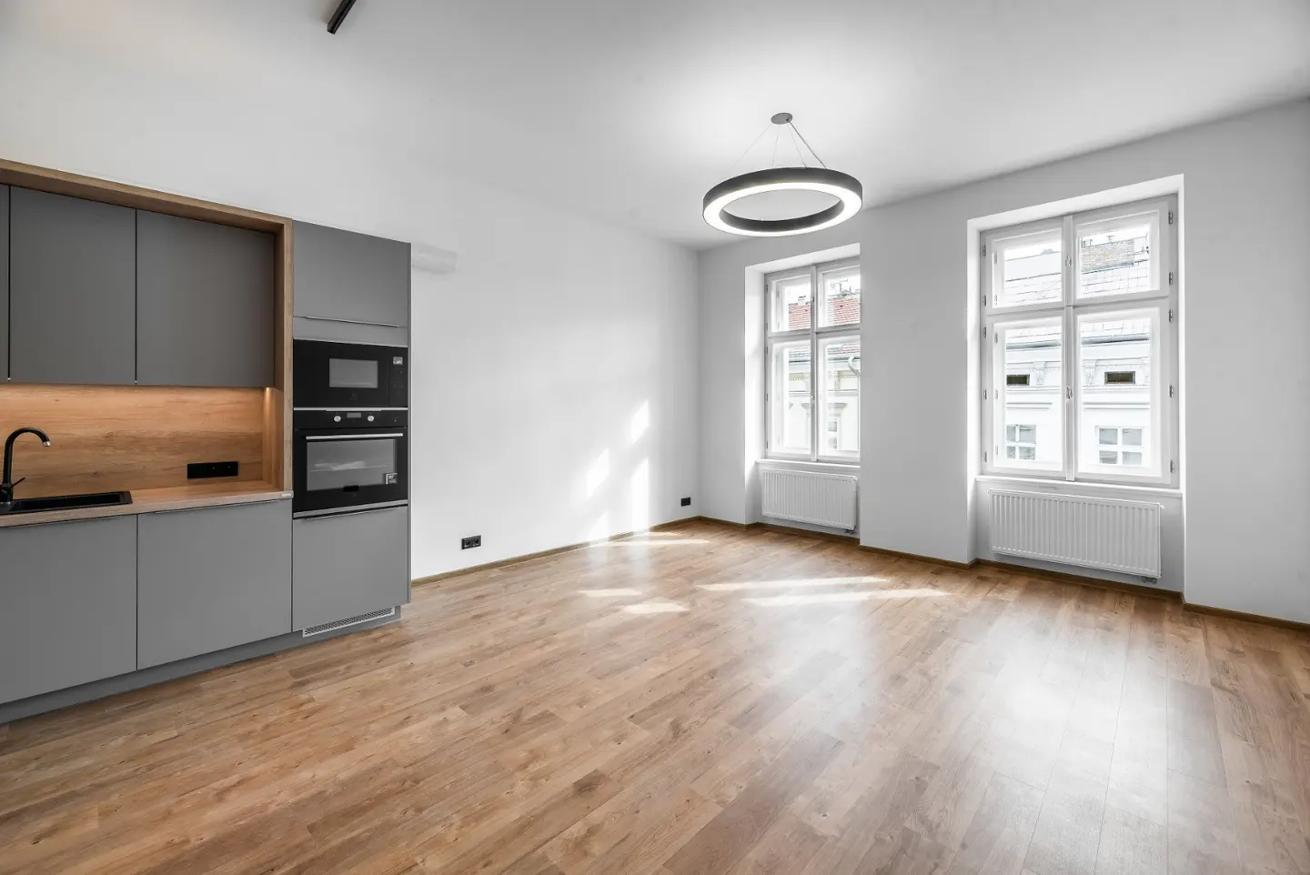 Bright, empty apartment with hardwood floors, white walls, and gray kitchen cabinets. Two large windows let in sunlight.