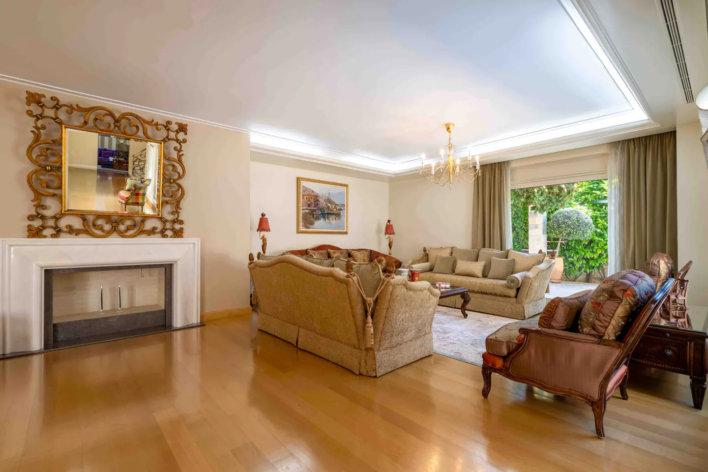 A living room with hardwood floors, beige walls, and ornate furniture. A fireplace with a gold-framed mirror sits on the left. Sofas and a chaise lounge face an open patio.