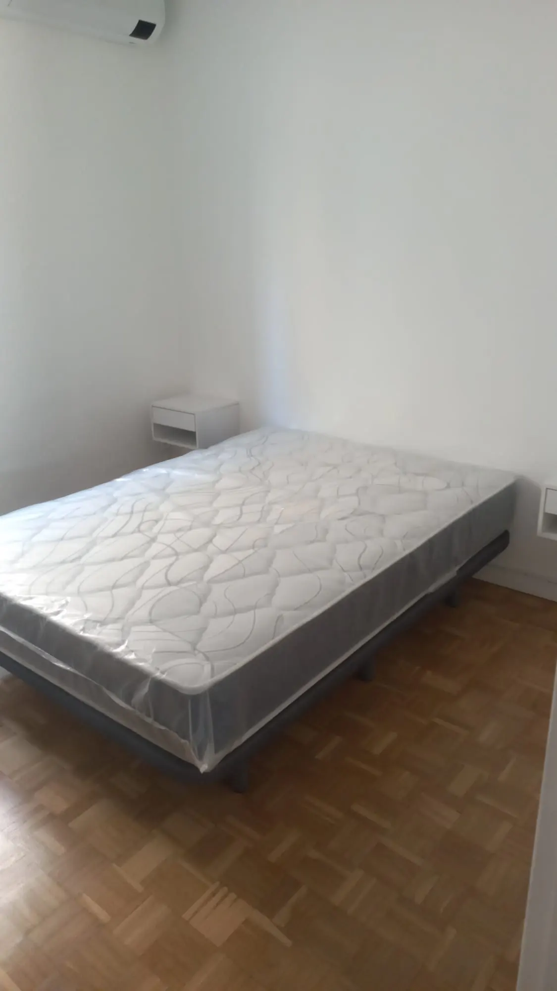 Bedroom with a mattress on a metal frame, a white nightstand, and hardwood floors against white walls.