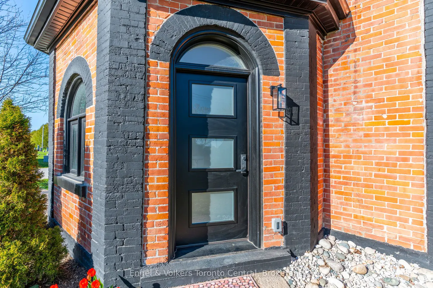 Exterior view of a brick building with a black door and trim, featuring an arched window and a modern light fixture.