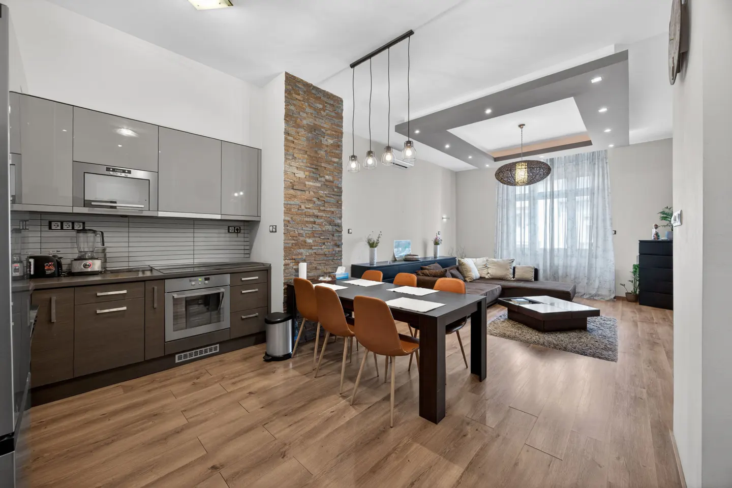 Modern open-plan kitchen and dining area with grey cabinets, a stone accent wall, and a living room with a brown sofa.