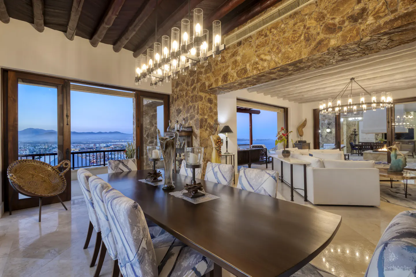 A dining room with a long wooden table, upholstered chairs, and a stone wall. Large windows offer a view of the city and ocean.