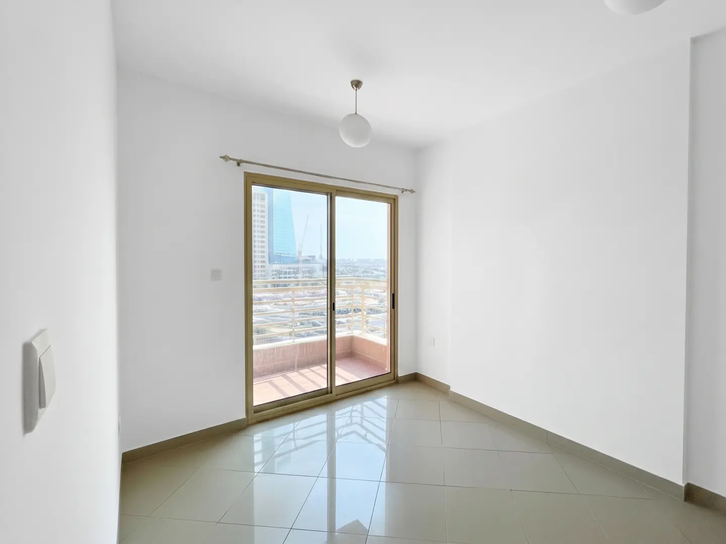 Bright, empty room with white walls, tile floor, and sliding glass doors to a balcony with a city view.