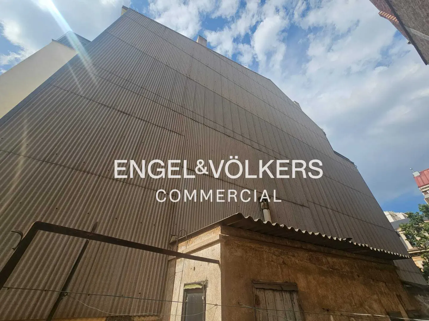 Low-angle view of a tall, corrugated metal building wall under a blue sky with the Engel & Völkers Commercial logo.