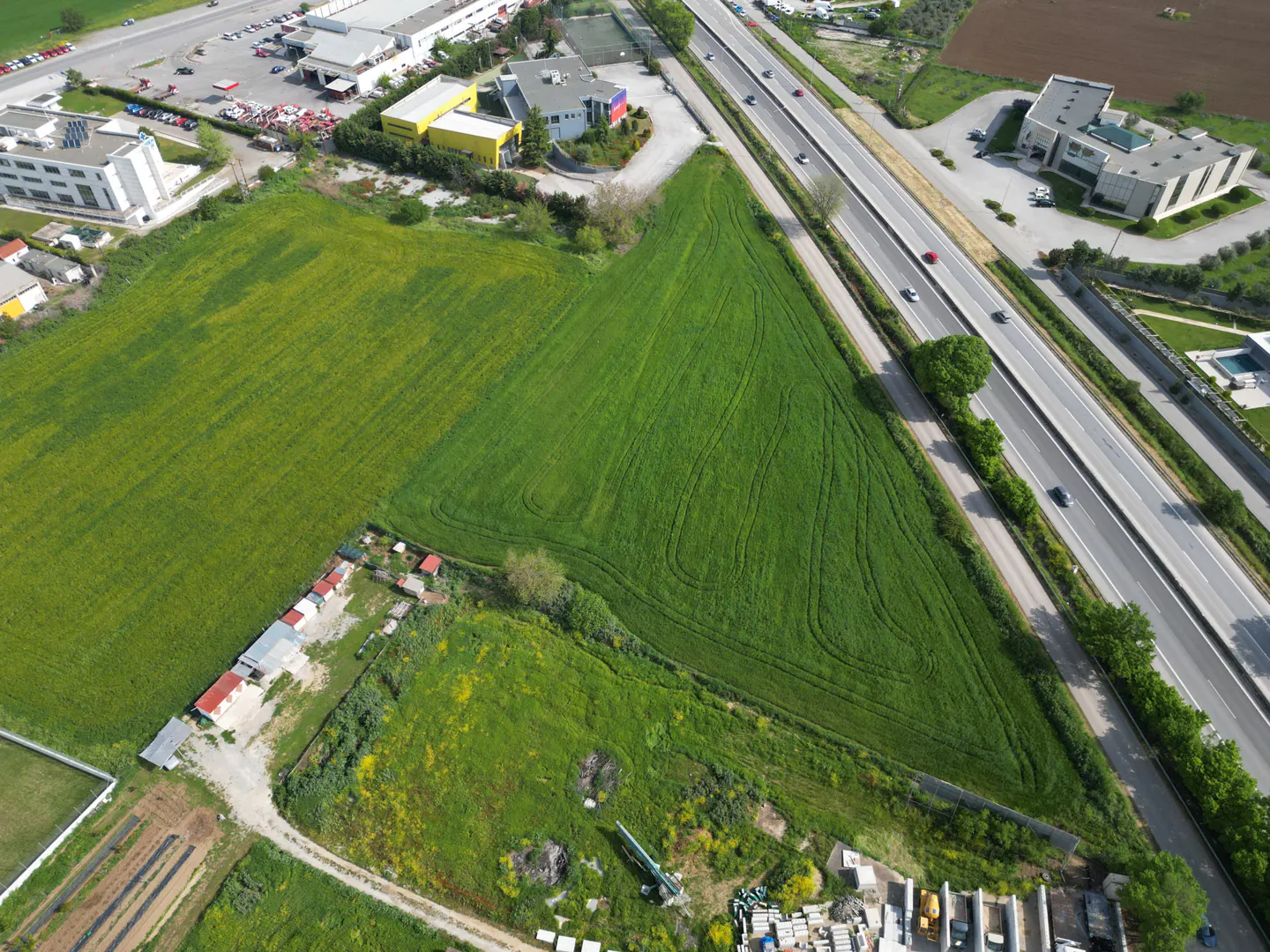 Aerial view of a green field next to a highway with cars, buildings, and trees.