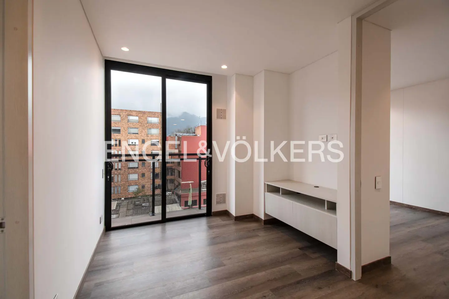 An empty room with dark wood floors, white walls, and a black-framed window with a balcony overlooking a city. The Engel & Völkers logo is superimposed on the window.