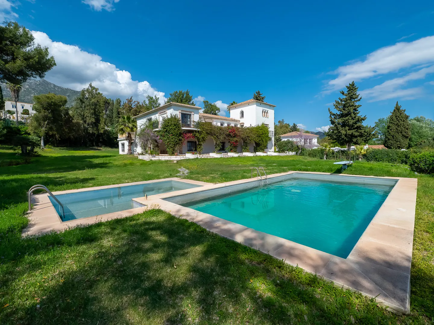 A white villa with a pool on a sunny day. The pool is turquoise and surrounded by green grass. The sky is blue with white clouds.