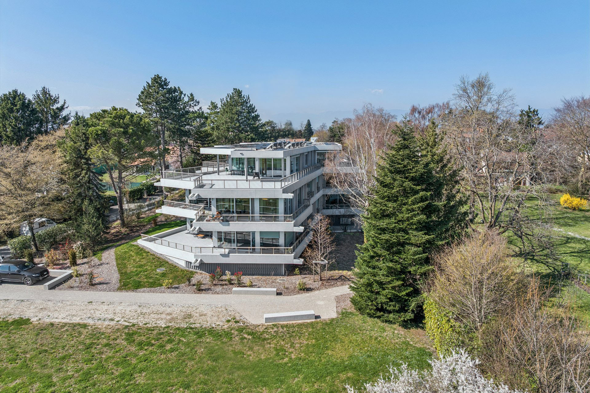 Modern, multi-story white building with large balconies, surrounded by green trees and grass under a clear blue sky.