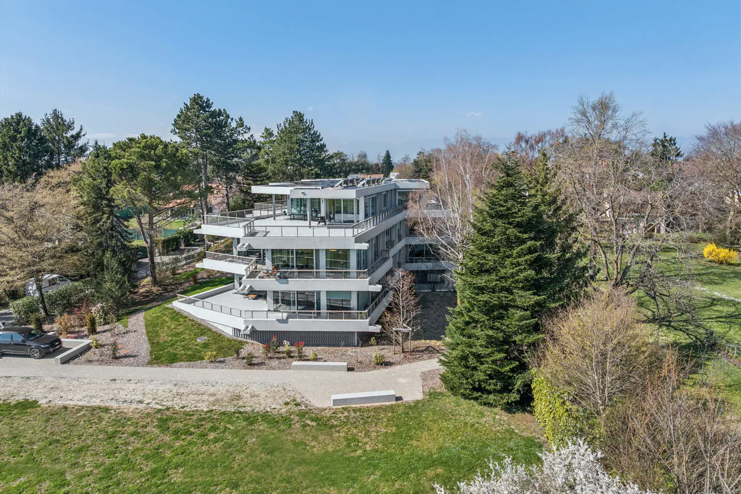 Modern, multi-story white building with large balconies, surrounded by green trees and grass under a clear blue sky.