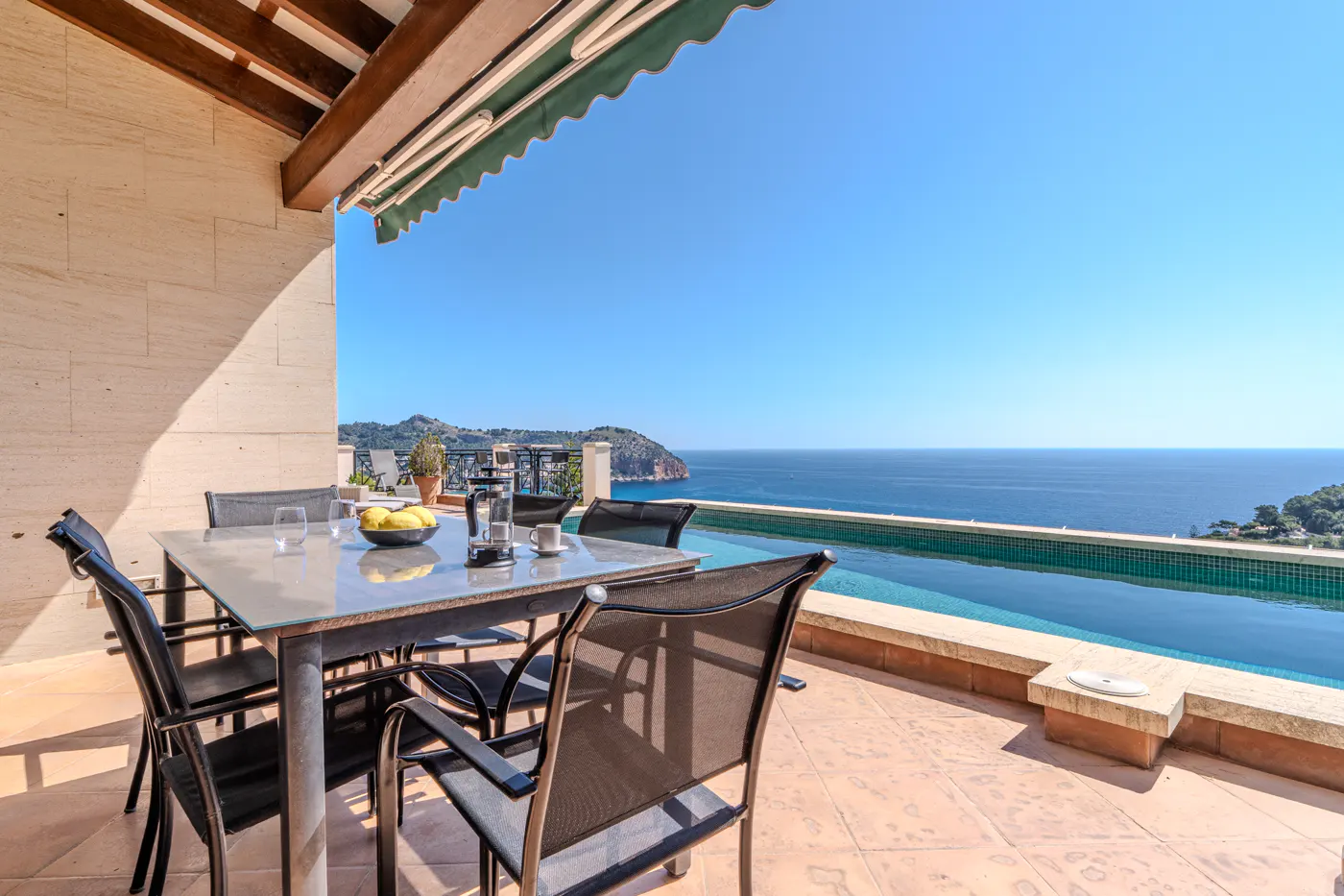 Outdoor patio with table, chairs, and infinity pool overlooking the ocean on a sunny day.