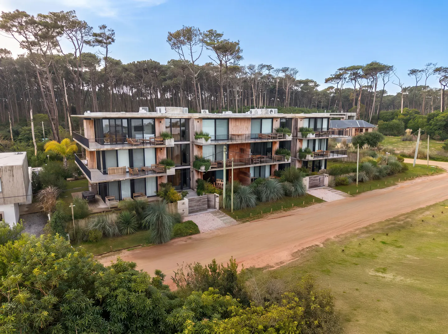 Modern three-story apartment building with wooden balconies and large windows, nestled in a forest near a dirt road.