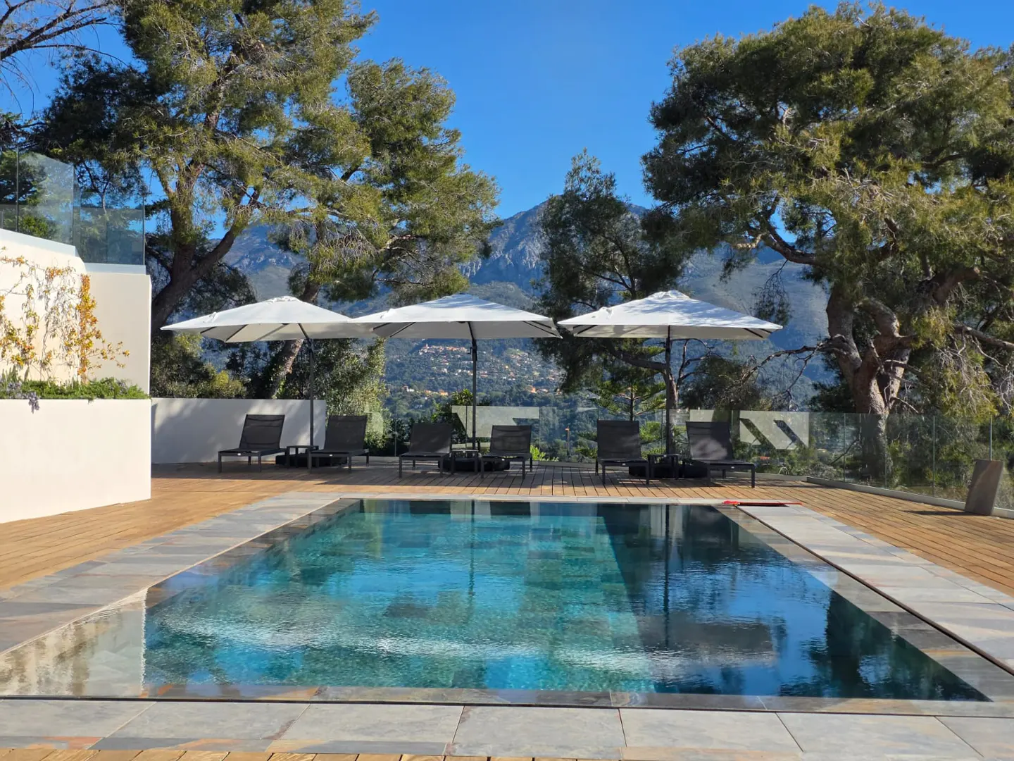 A rectangular pool with lounge chairs and umbrellas on a wooden deck, with a mountain view.