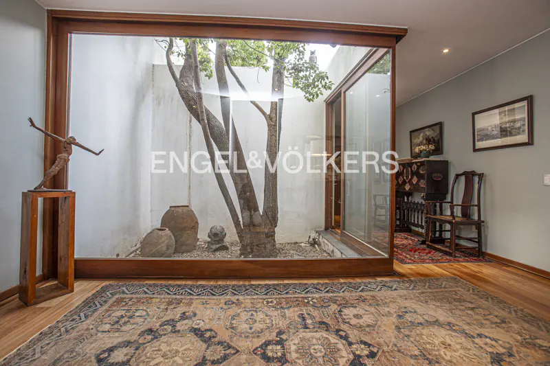 Interior view of a room with hardwood floors, a patterned rug, and a glass-enclosed courtyard with a tree and pottery. A bronze statue stands on a wooden pedestal.