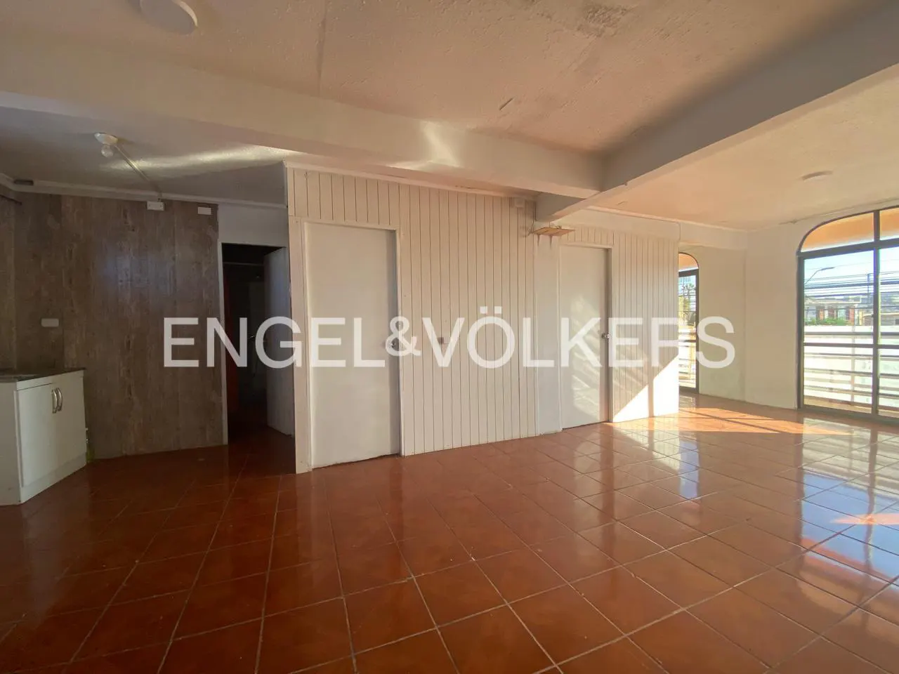 Empty room with brown tile floor, white walls, and two white doors. Large windows let in sunlight. Engel & Völkers logo is superimposed.