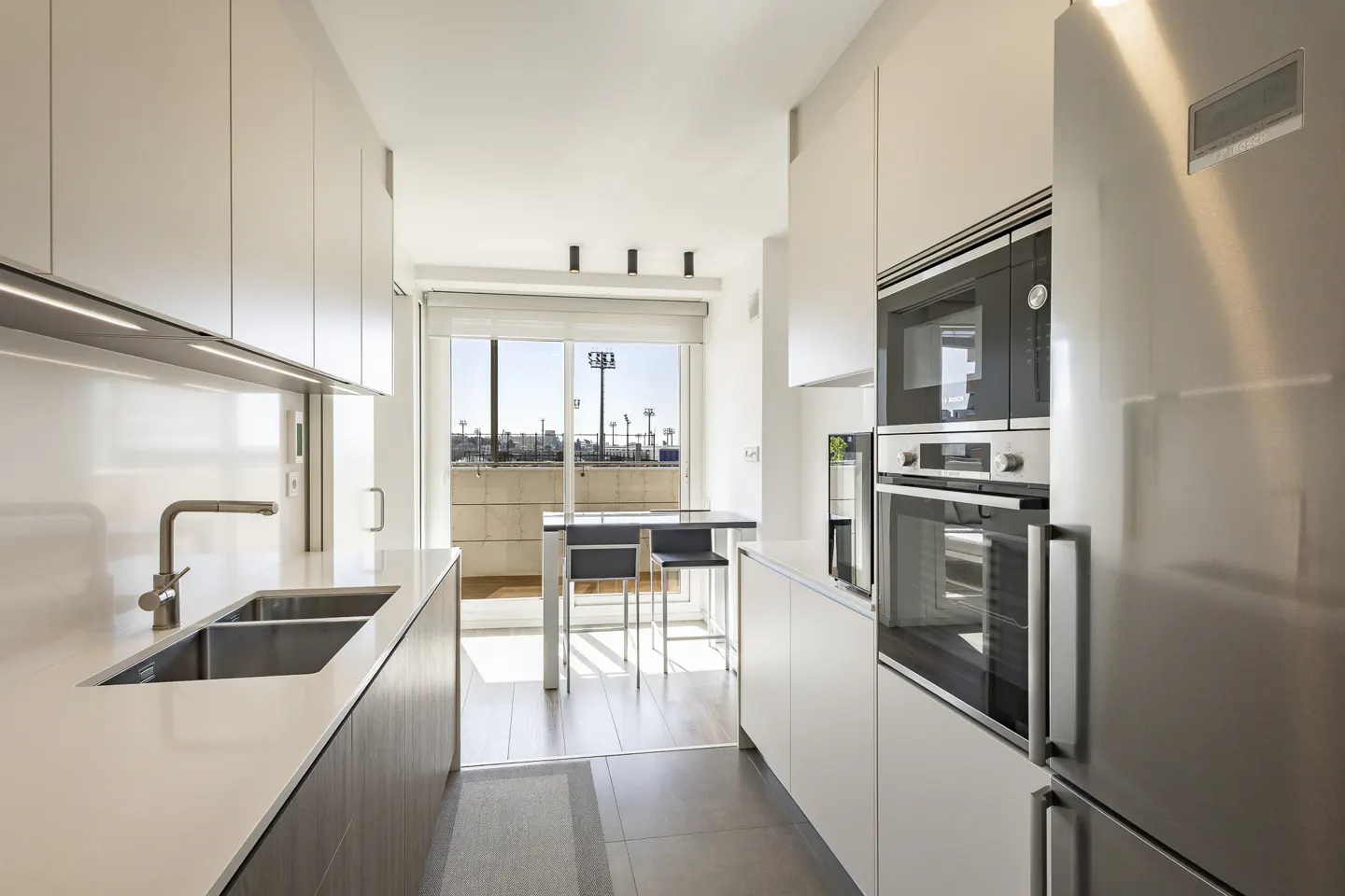 Bright, modern kitchen with white cabinets, stainless steel appliances, and a sink. A table and chairs are visible through a sliding glass door.