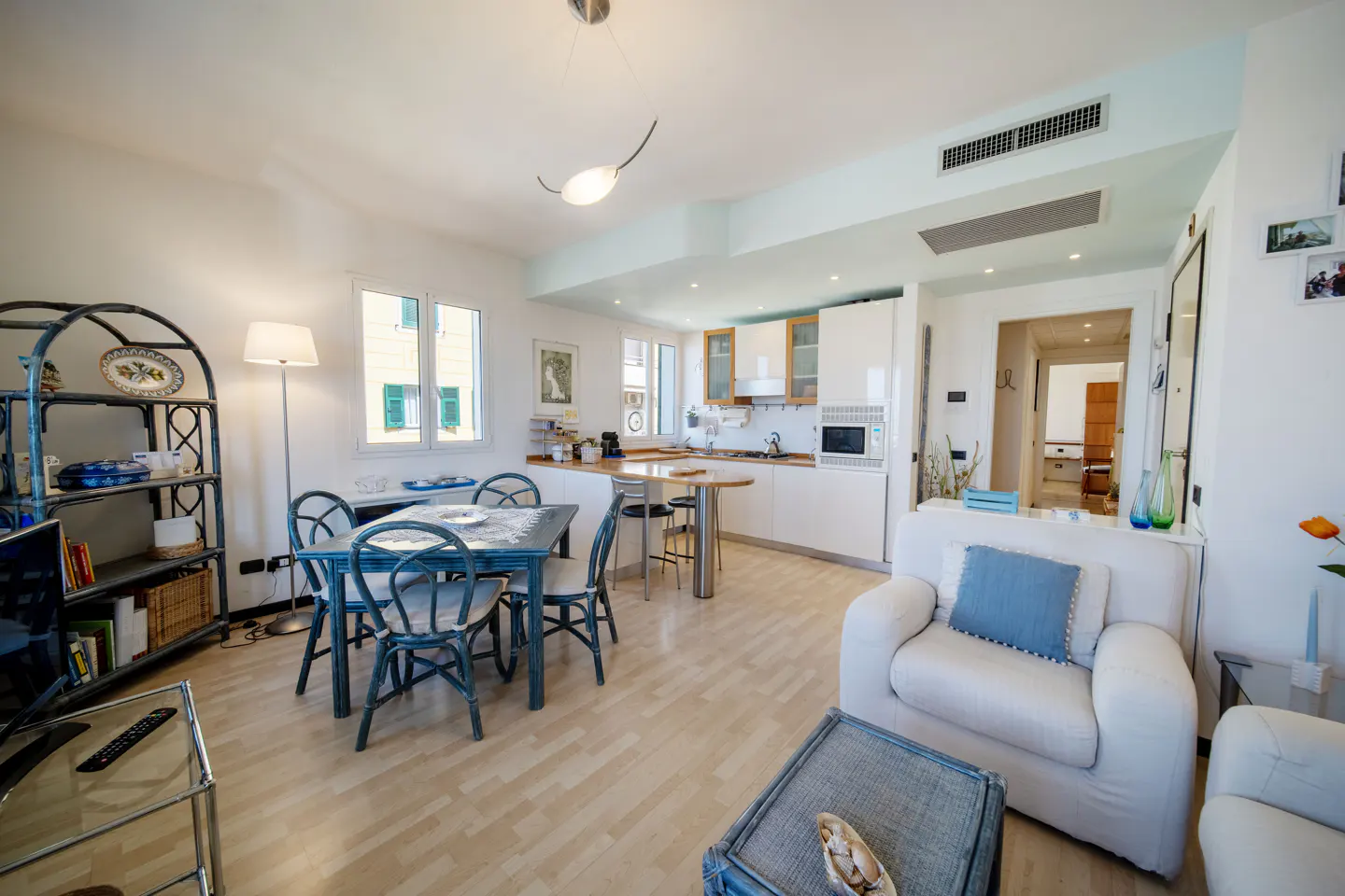 Open-plan living area with a blue dining set, white kitchen, and a comfortable white armchair with a blue cushion.
