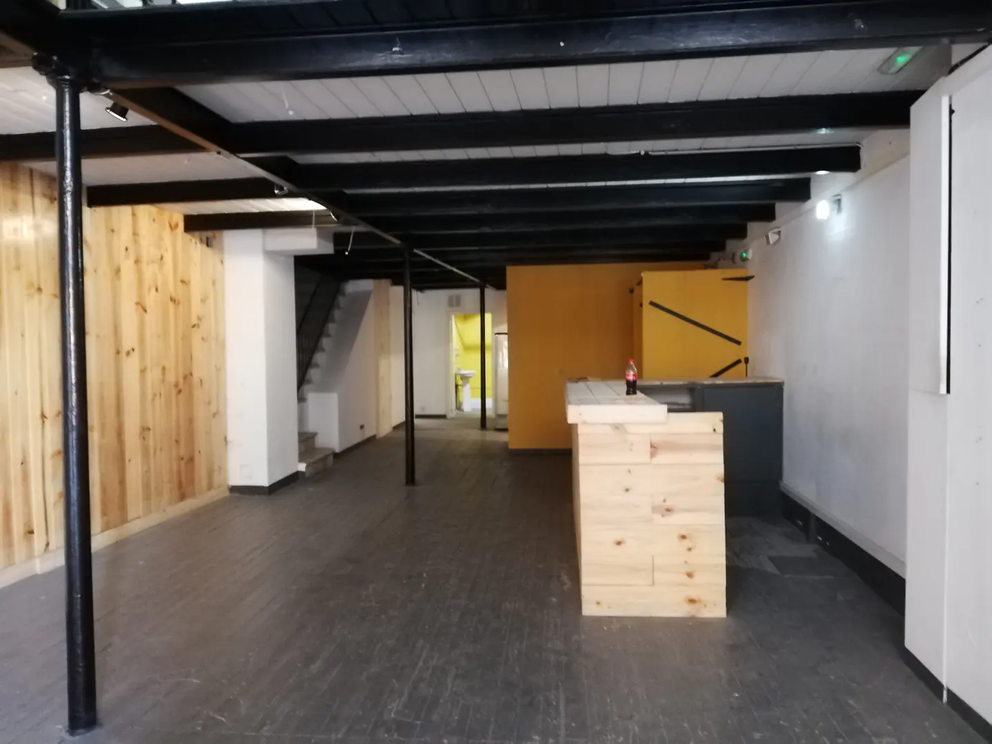 An empty commercial space with wood walls, black ceiling beams, and a wooden bar. Stairs lead to an upper level.