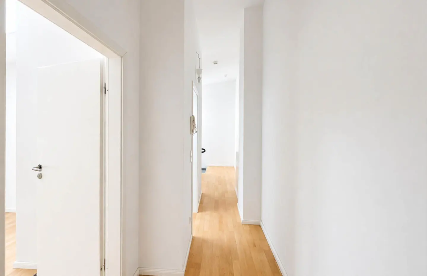 Bright, narrow hallway with light wood floors and white walls. An open white door is on the left.