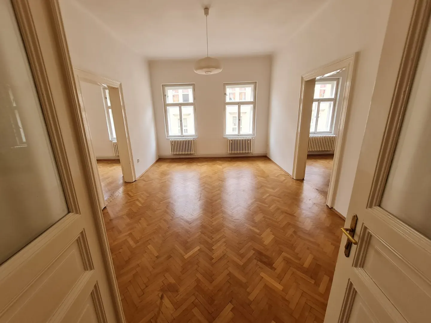 Empty room with white walls, herringbone wood floors, two windows with radiators, and doorways leading to adjacent rooms.
