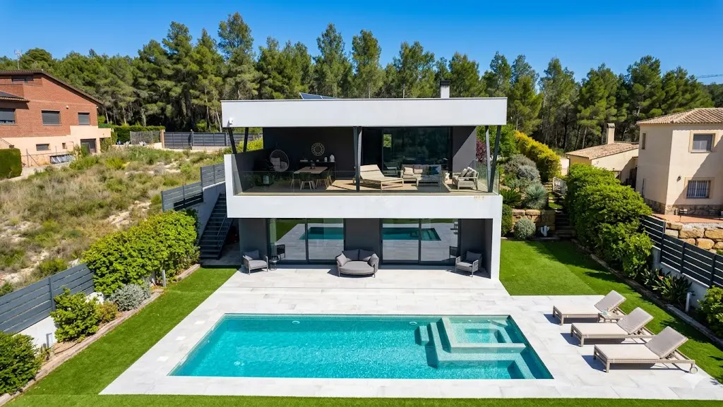 Two-story modern house with a pool, patio furniture, and a balcony overlooking the backyard. Trees in the background.