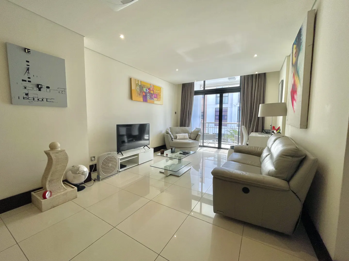 Bright living room with beige leather sofa, round chair, glass table, and TV. Balcony visible through sliding glass doors. Artwork on walls.