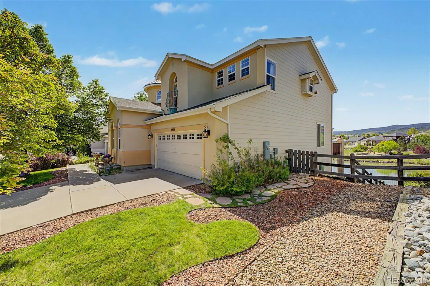 Two-story beige house with a two-car garage, concrete driveway, green lawn, and a gravel yard beside a small pond.