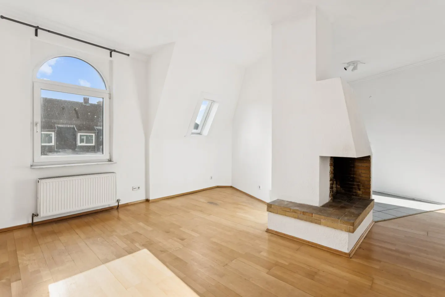Bright, empty room with hardwood floors, white walls, and a brick fireplace. A large arched window shows a view of rooftops.
