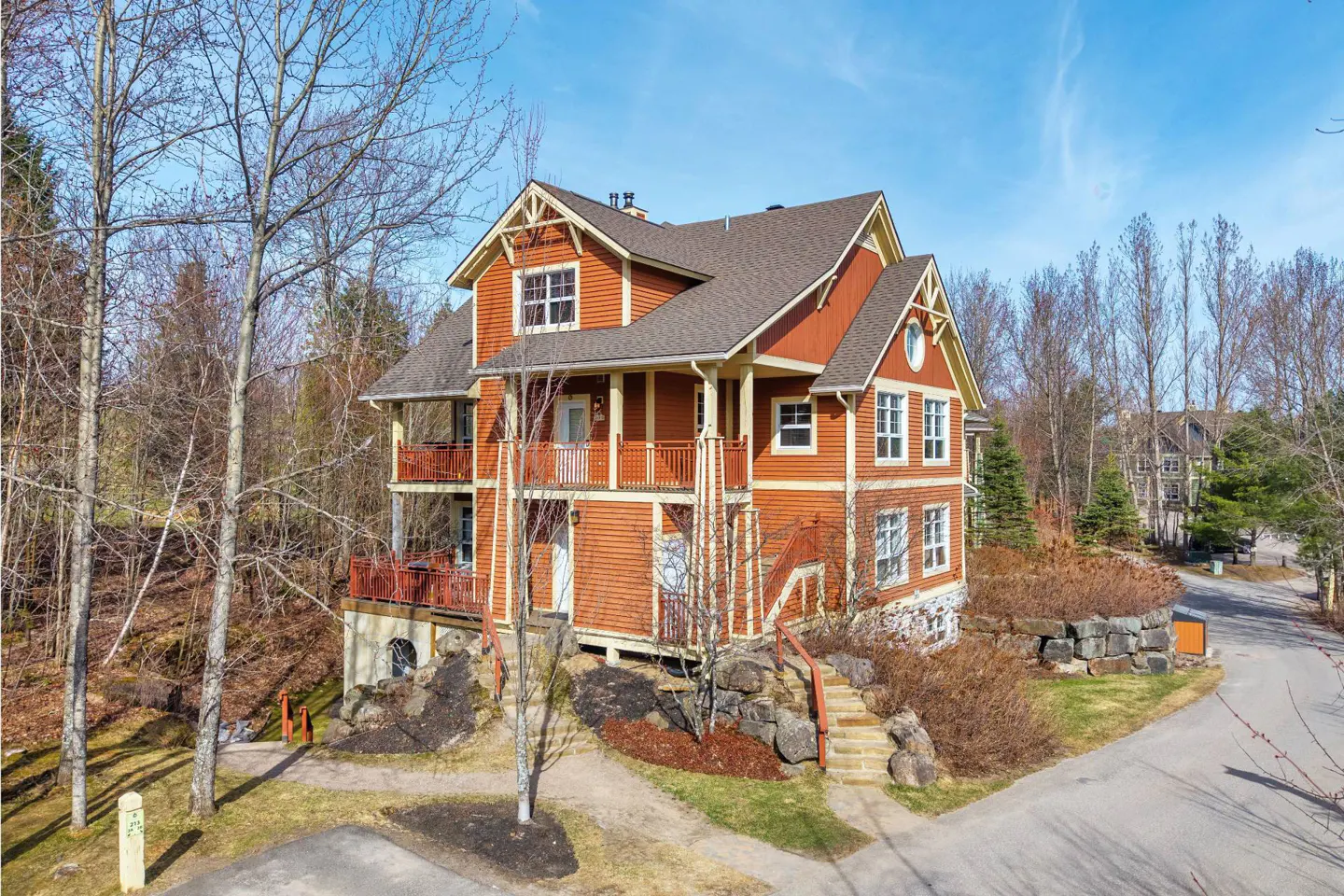 Two-story orange house with brown roof, surrounded by trees and a stone path leading to the entrance. Balconies on both floors. Blue sky.