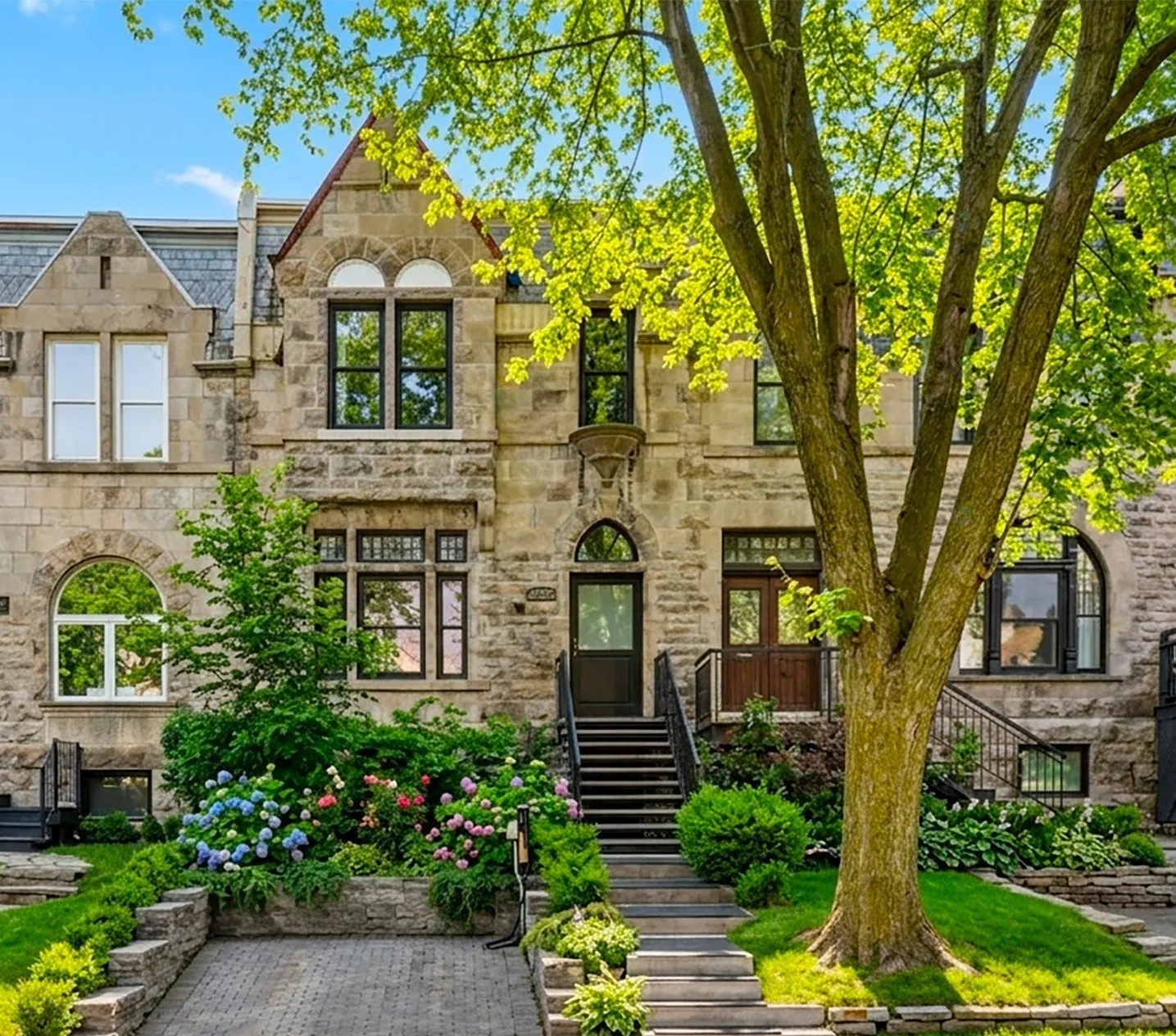 Stone townhouse with black windows and door, surrounded by green trees and colorful flowers. Steps lead to the entrance.