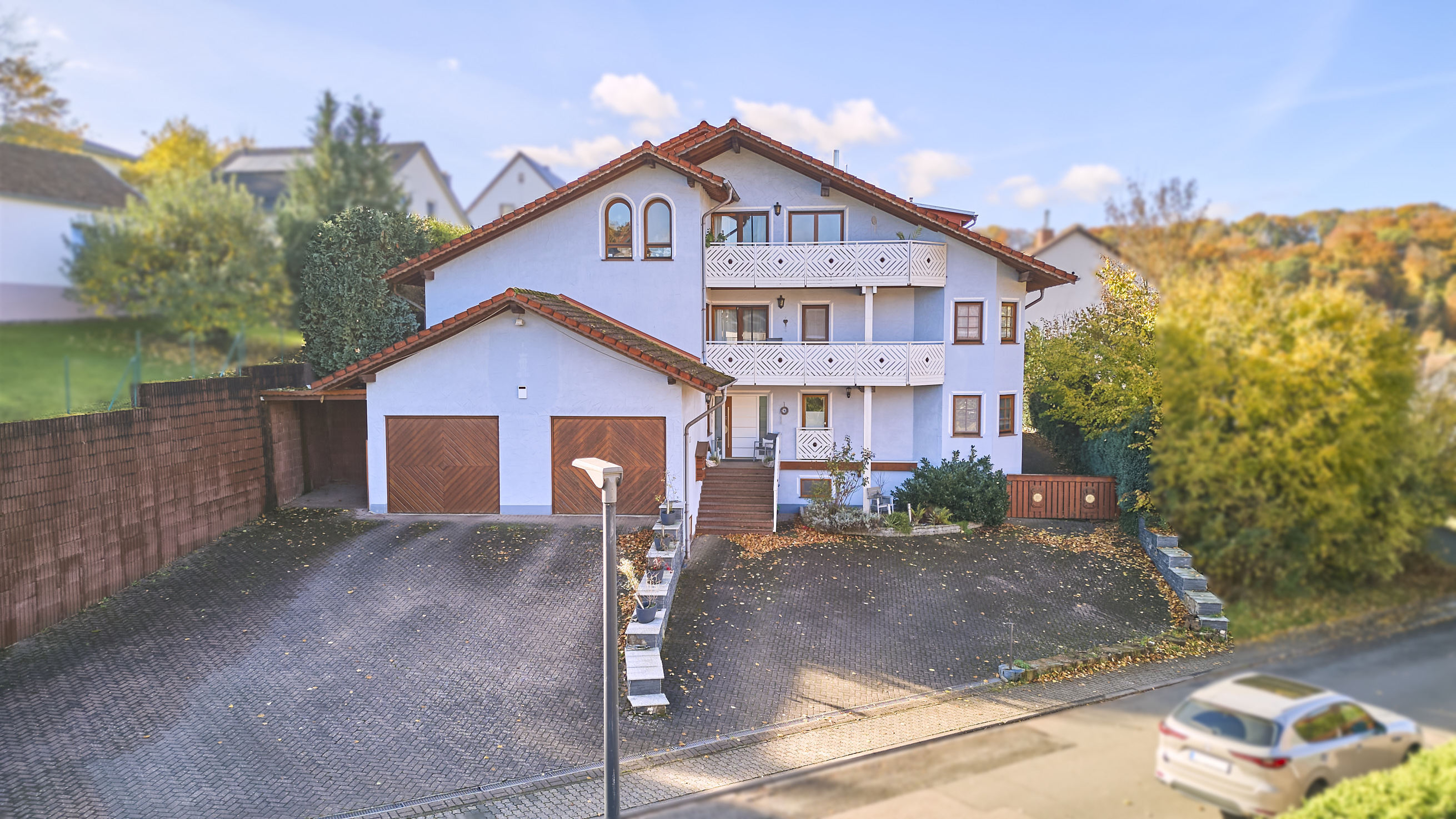 Two-story light blue house with red roof, two balconies, and attached garage with brown doors. A car is parked on the street. [AI-generated text]