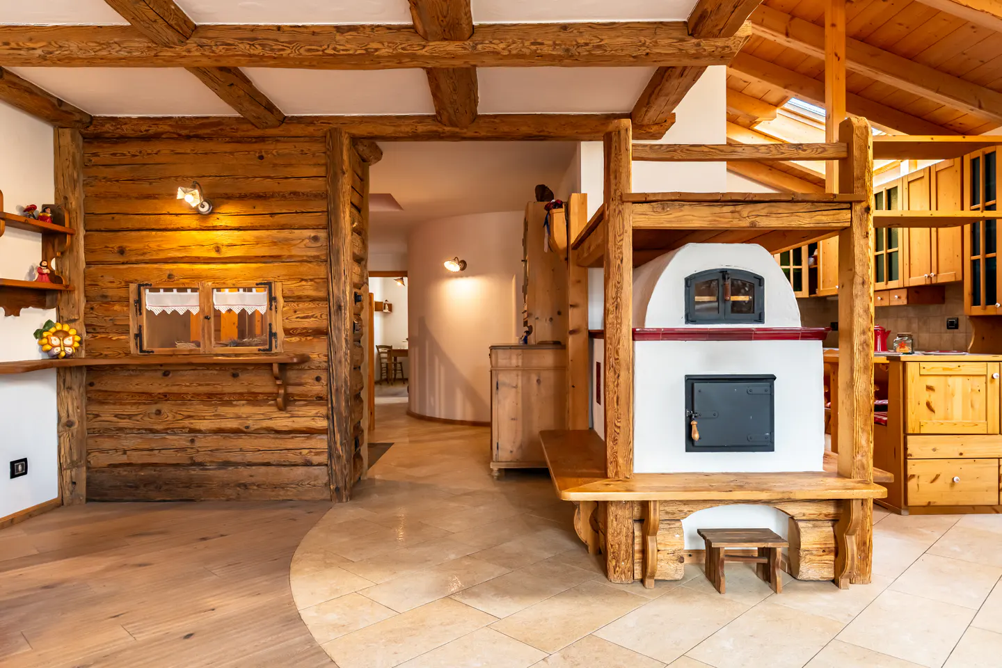 Rustic interior with log walls, a white masonry stove, wooden beams, and a transition from wood to tiled flooring.