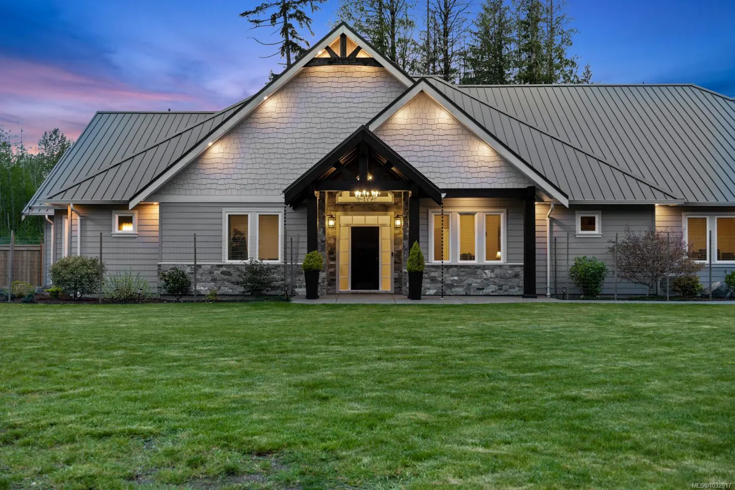 A gray house with a metal roof and a green lawn at dusk. The house has a black door and a stone foundation.
